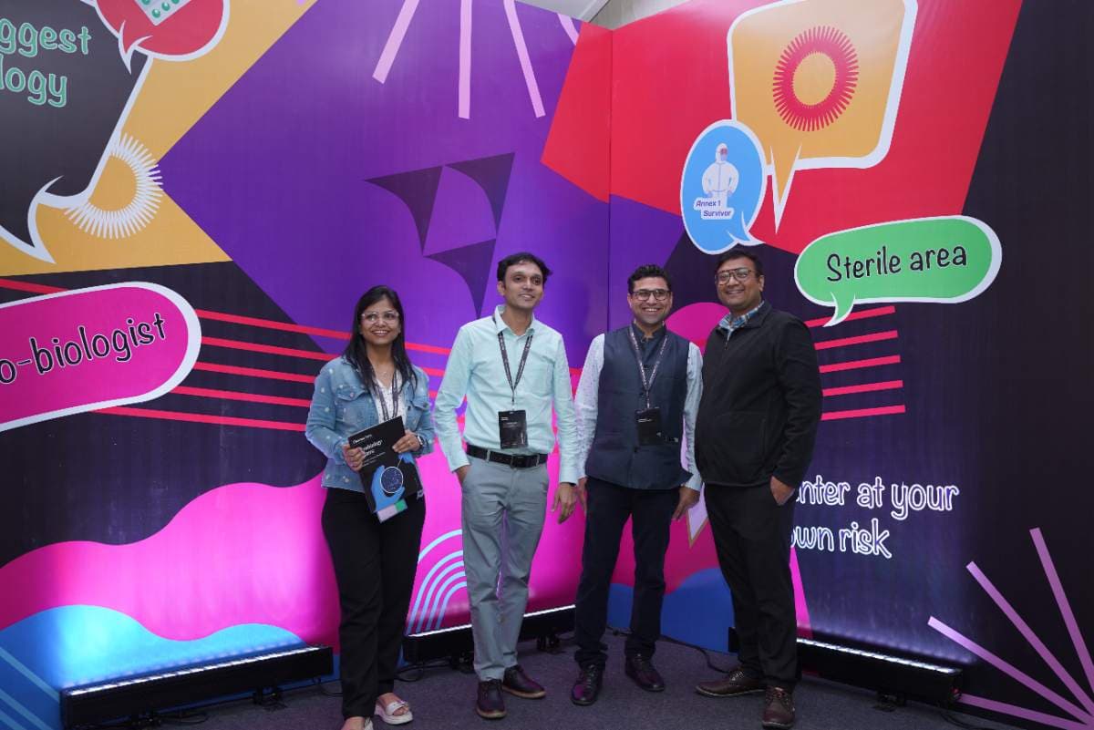 Four delegates posing in front of the illustrated microbiology wall at Pharmig India 2025